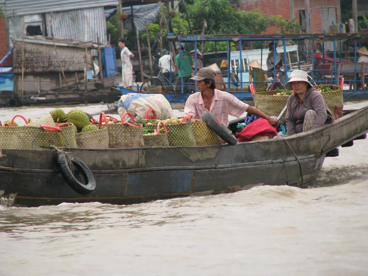 Thuyền gỗ chở đầy sọt sầu riêng và trái cây, hai người ngồi trên thuyền tại một khu chợ trên sông