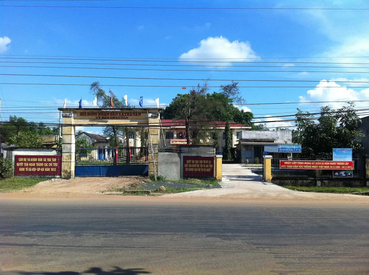 Cổng trụ sở uỷ ban nhân dân xã với hàng rào, cây xanh và các bảng thông tin đỏ hai bên