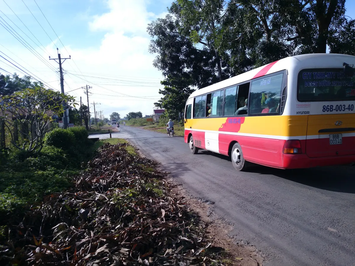 Một xe khách giường nằm chạy trên đường nhựa hai chiều, ven đường là cây xanh và vỏ hạt phơi trên lề
