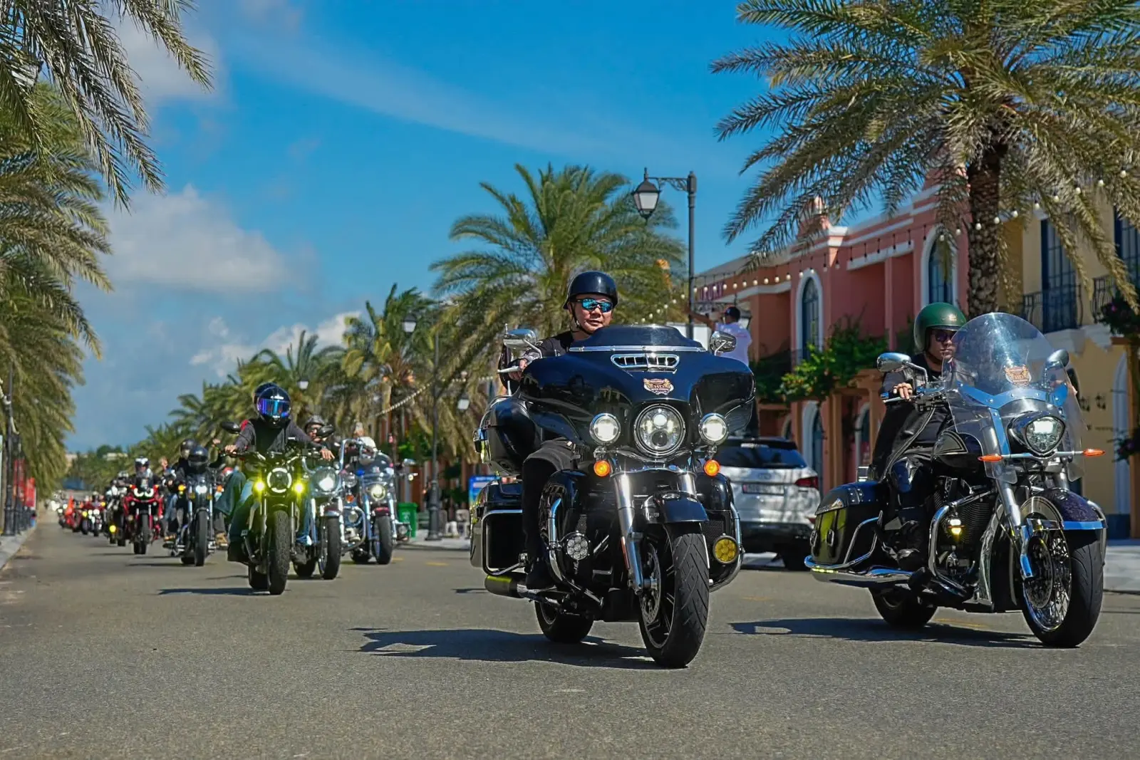 Ba mô tô Harley-Davidson chạy qua dãy phố kiến trúc châu Âu, hai bên đường trồng dừa, phía sau là đoàn xe nối dài