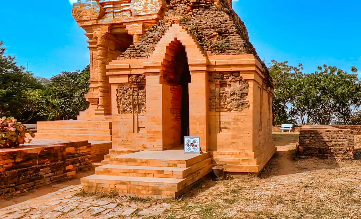 Một tháp Chăm bằng gạch với cửa vòm nhọn, bậc đá dẫn vào, tường móng gạch của công trình cũ hai bên